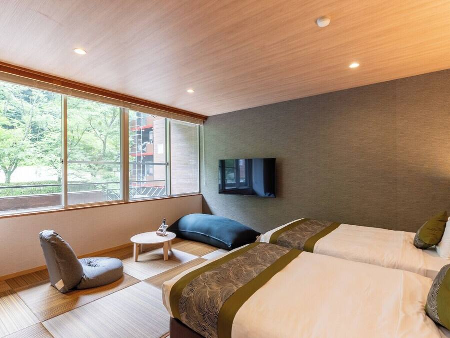 A hotel room with two beds, a TV mounted on the wall, a small table, and a window with a view of trees.