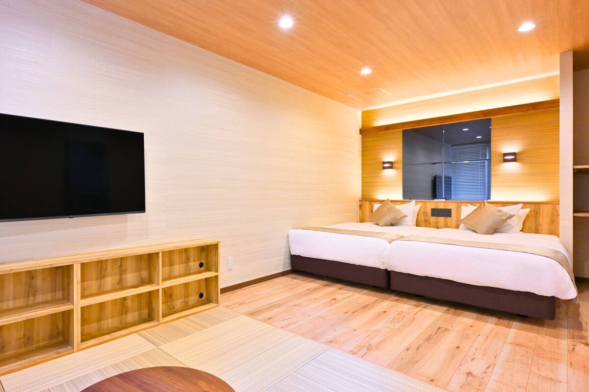 A modern bedroom with a large flat-screen TV mounted on the wall, a bed with a wooden headboard, and built-in wooden shelving below.
