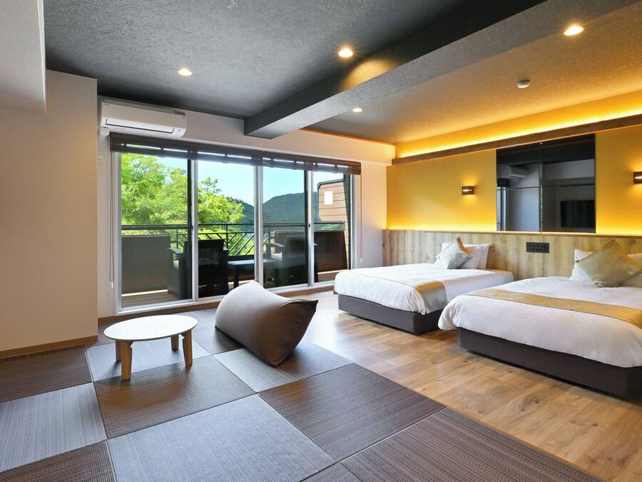 Habitación de hotel moderna con dos camas, una mesita pequeña y un amplio ventanal corredero que da a un espacio exterior.
