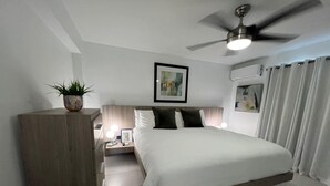 Una habitación de hotel moderna con una cama grande, una mesita de noche con lámpara, un ventilador de techo y una planta en maceta.