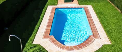 Piscina rectangular con borde de azulejos, rodeada por un césped cuidado y un patio con sillas de descanso y una mesa.