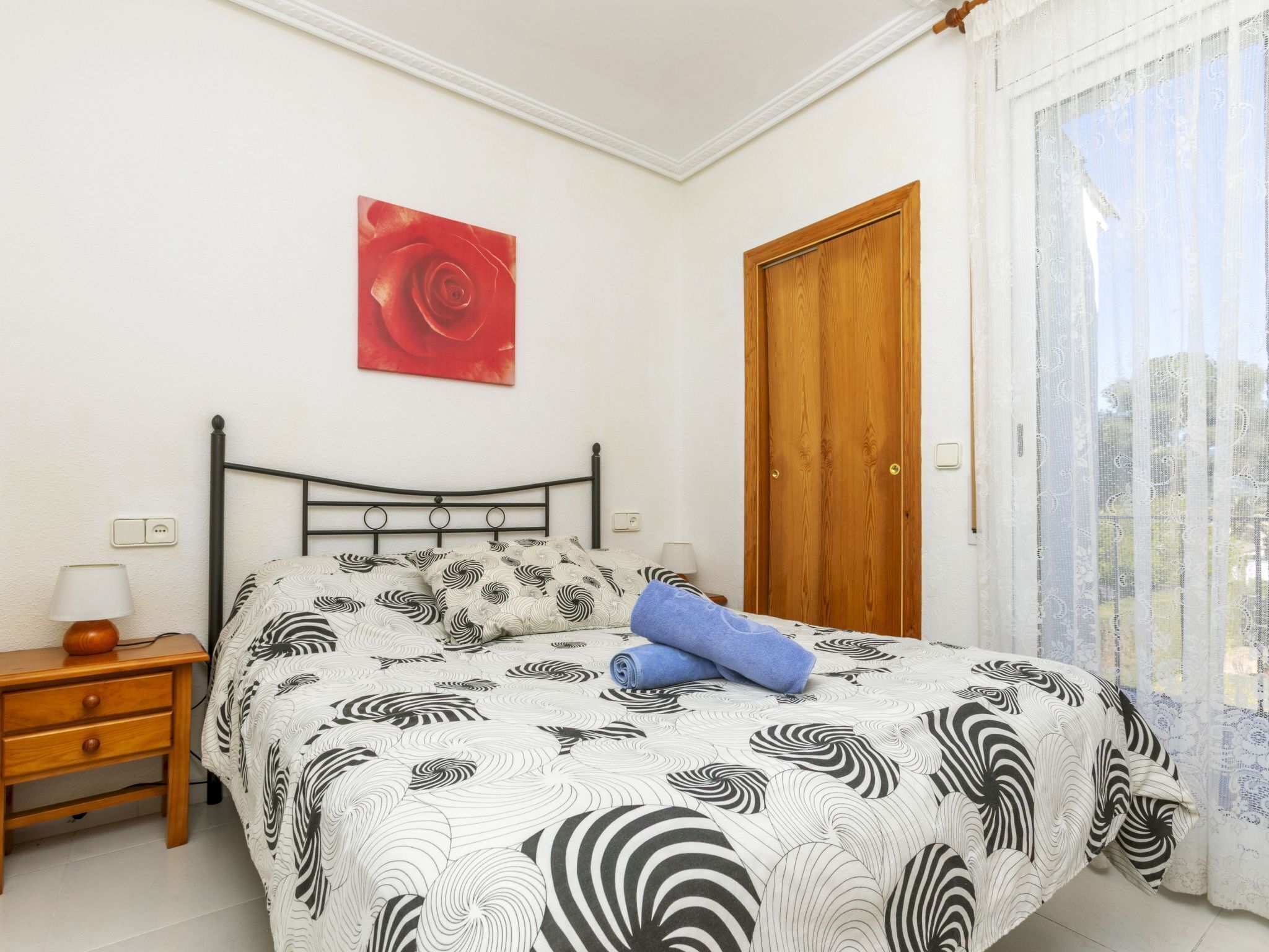 A bedroom with a bed, a nightstand, a wooden door, and a window with sheer curtains.