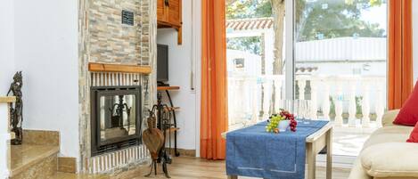A living room with a fireplace, a table with fruit, and a balcony with a view of trees.