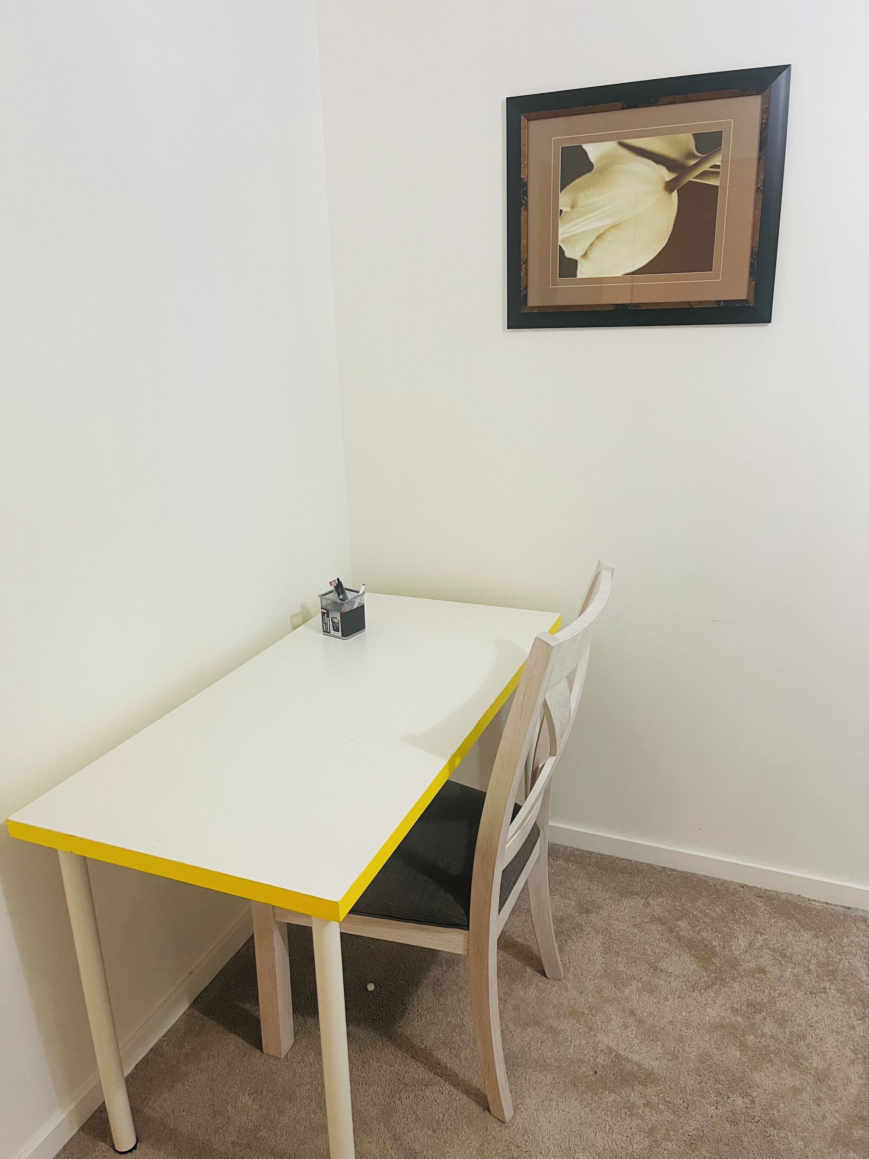 A small, white table with a yellow edge, a chair with a black cushion, and a framed picture on the wall.