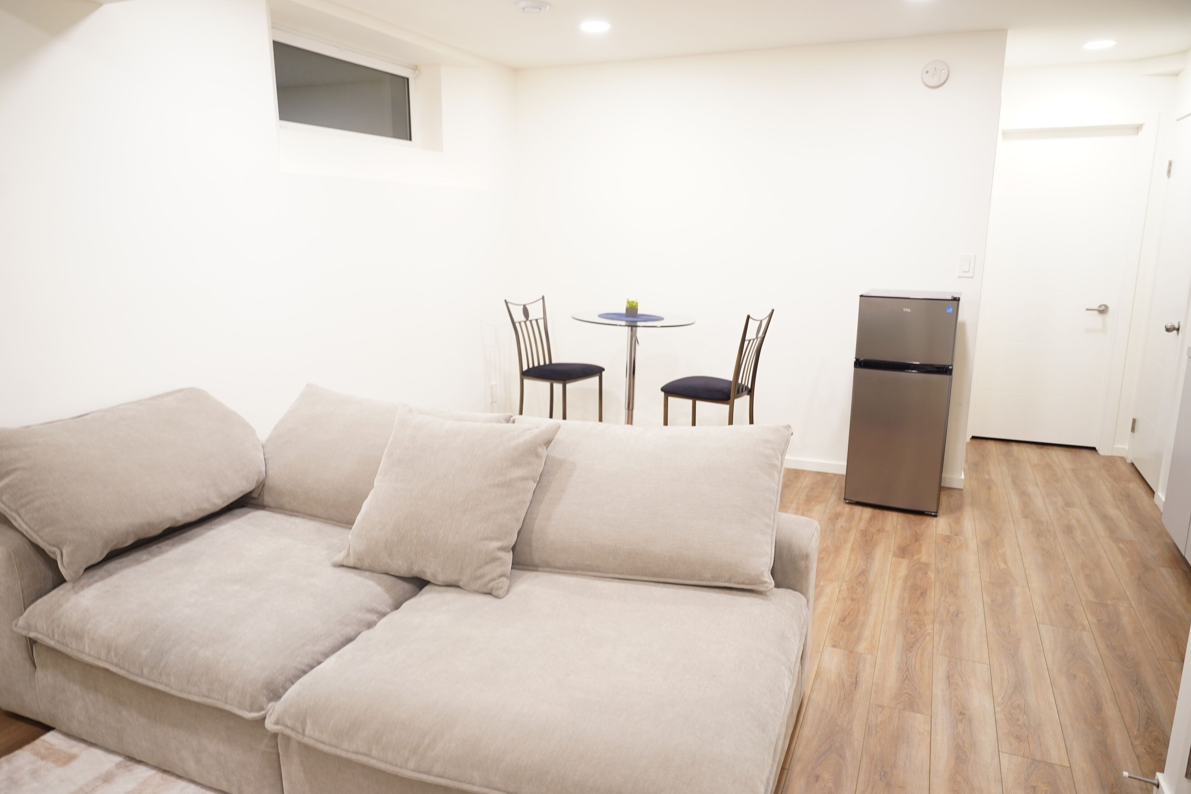 A compact living space with a beige sofa, a small round table with two chairs, a built-in refrigerator, and a white door.