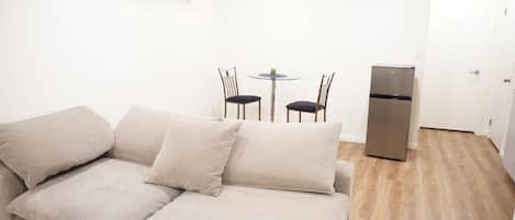 A compact living space with a beige sofa, a small round table with two chairs, a built-in refrigerator, and a white door.
