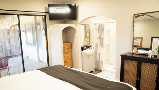 A bedroom with a bed, a TV mounted on the wall, a bathroom with a sink and mirror, and a wooden dresser.