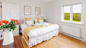 A bedroom with a large bed, white walls, a window with a view of the countryside, and a vase of flowers on a wooden table.