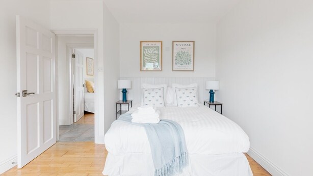 A bedroom with a large bed, two bedside tables with lamps, a door leading to another room, and two framed artworks on the wall.