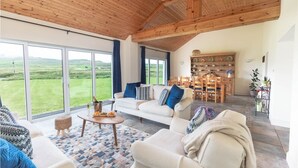 A spacious living area with a high ceiling, large windows, a sofa set, a dining table, and a bookshelf.
