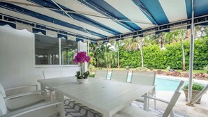 Un patio techado con una mesa y sillas blancas, una planta pequeña en maceta y una piscina al fondo.