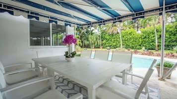 Eine überdachte Terrasse mit weißem Tisch und Stühlen, einem kleinen Topfpflanzen und einem Pool im Hintergrund.