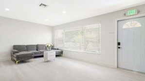 Una habitación amplia con un sofá gris, una mesita auxiliar blanca y una ventana con persianas. Se ve una señal de salida sobre una puerta.