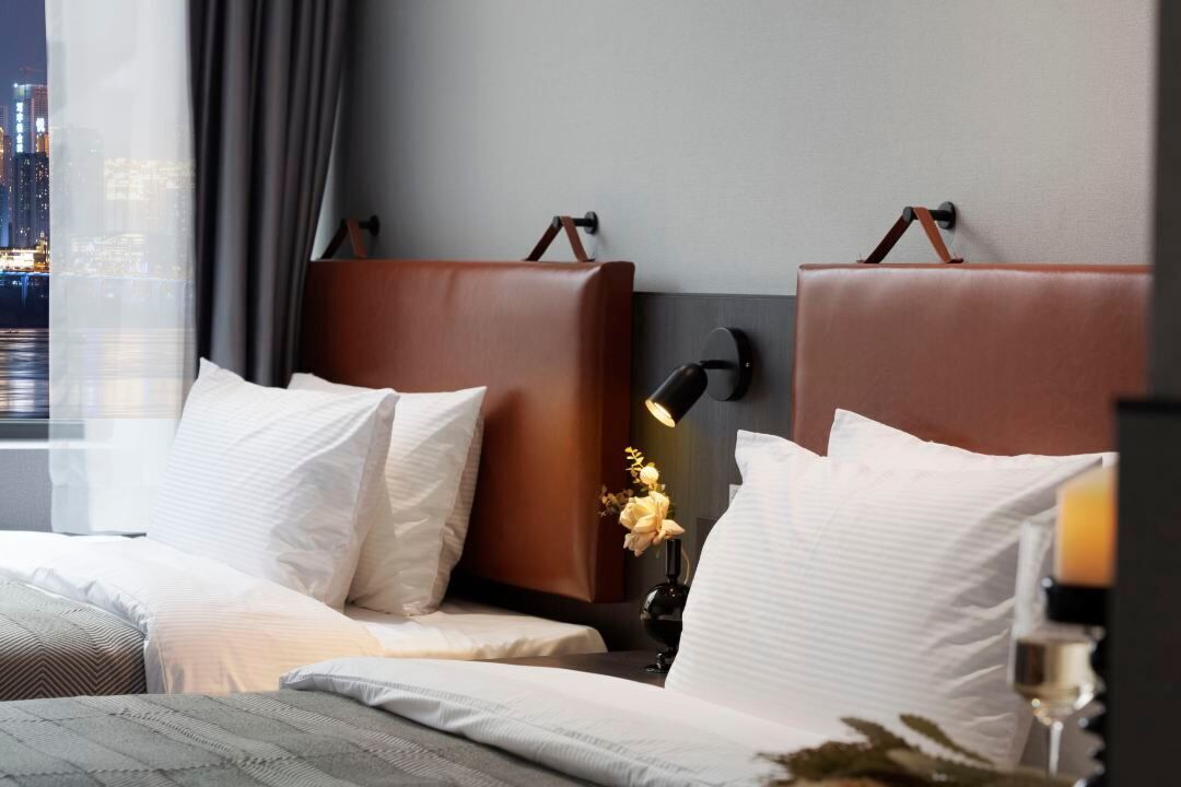 Una habitación de hotel moderna con vistas a la ciudad, una cama con sábanas blancas, un cabecero de cuero marrón y una mesita de noche con una lámpara y flores.