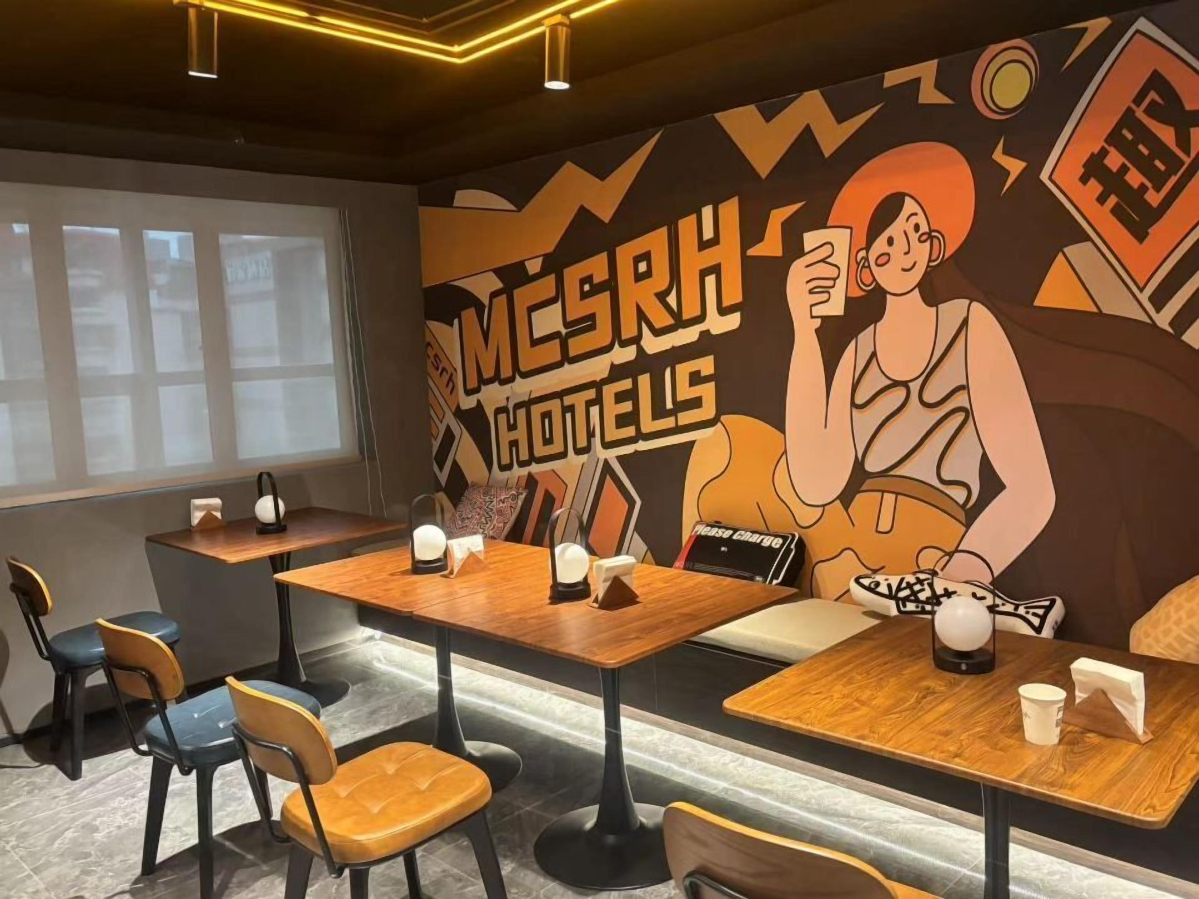 Interior de una cafetería moderna con mesas y sillas de madera, un mural con un personaje de caricatura y un cartel que dice 'Por favor, cargue' sobre las mesas.