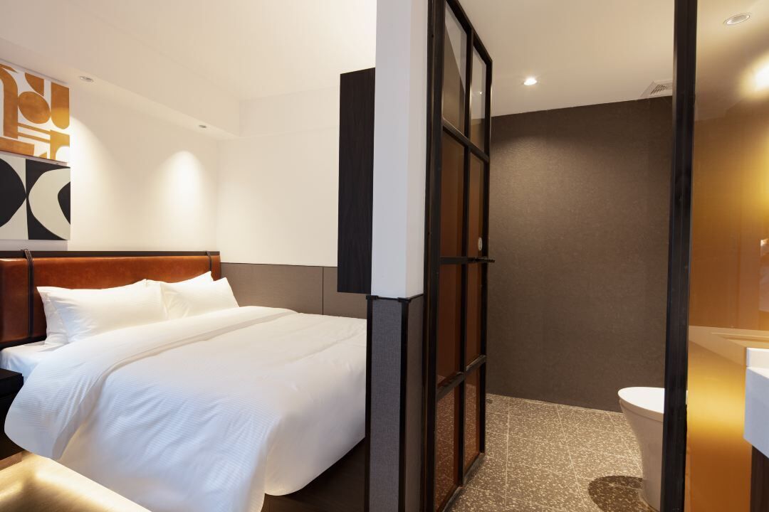 Una habitación de hotel con una cama grande, un cabecero de madera, un baño moderno con mampara de vidrio y una obra de arte colgada en la pared.
