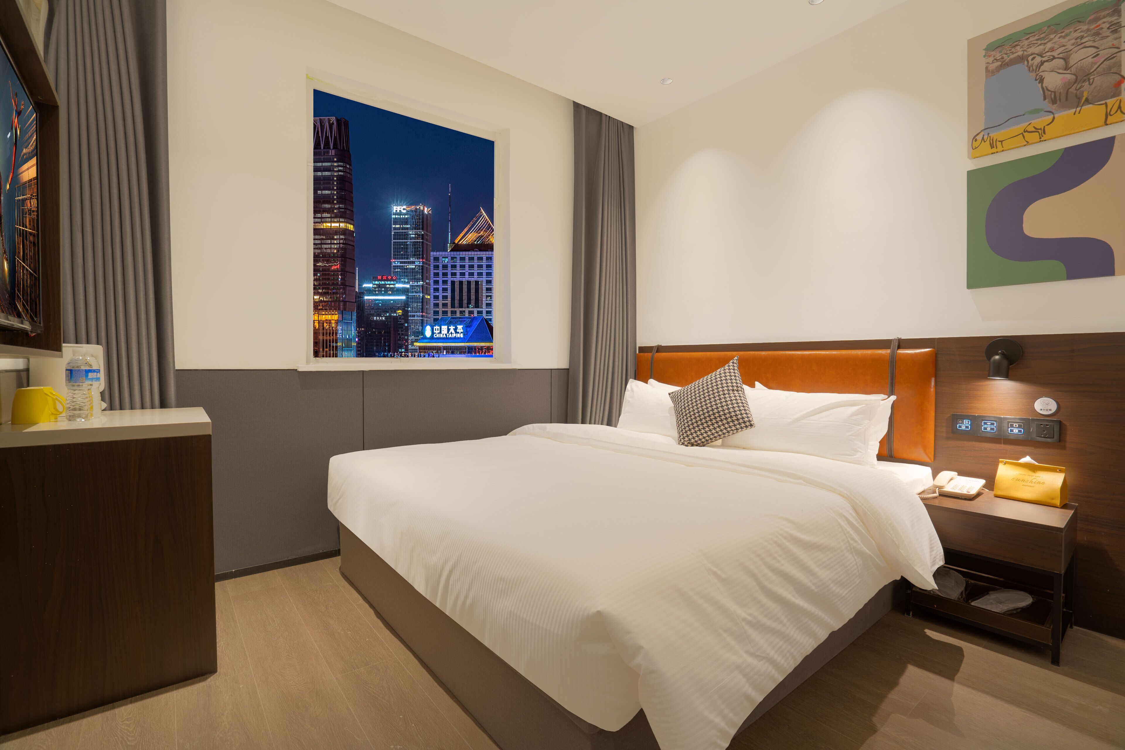 Habitación de hotel con una cama grande, vista nocturna de la ciudad, un escritorio con teléfono y una mesita de noche con lámpara.
