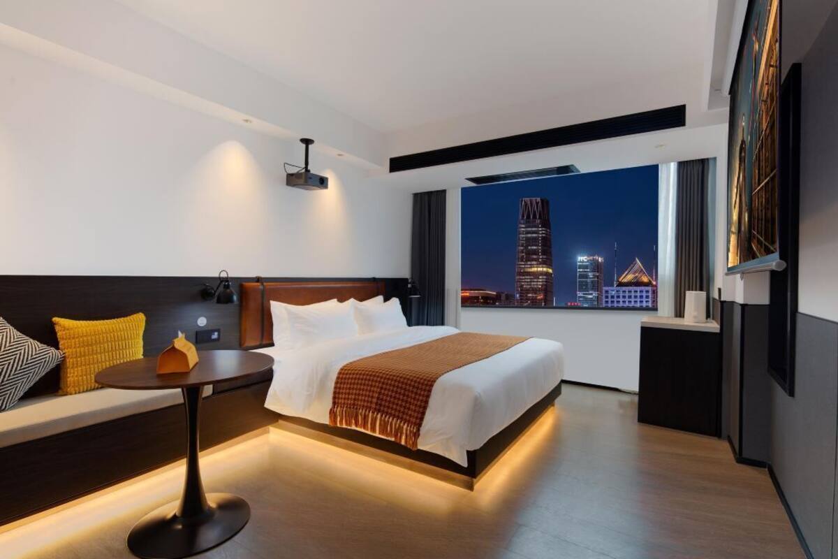 A modern hotel room with a large bed, a seating area with a table, and a view of a cityscape at night.