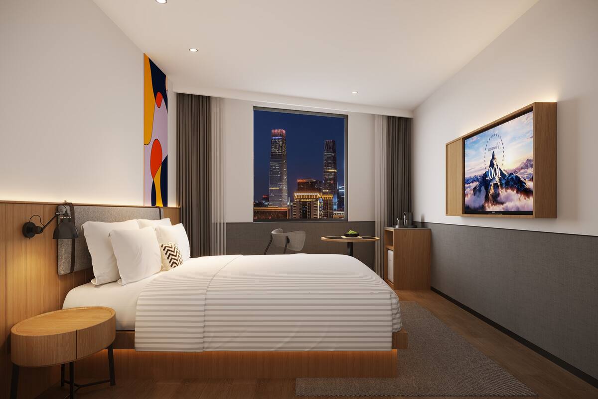 A modern hotel room with a large bed, a desk, a chair, a small table, and a large window with a city view.