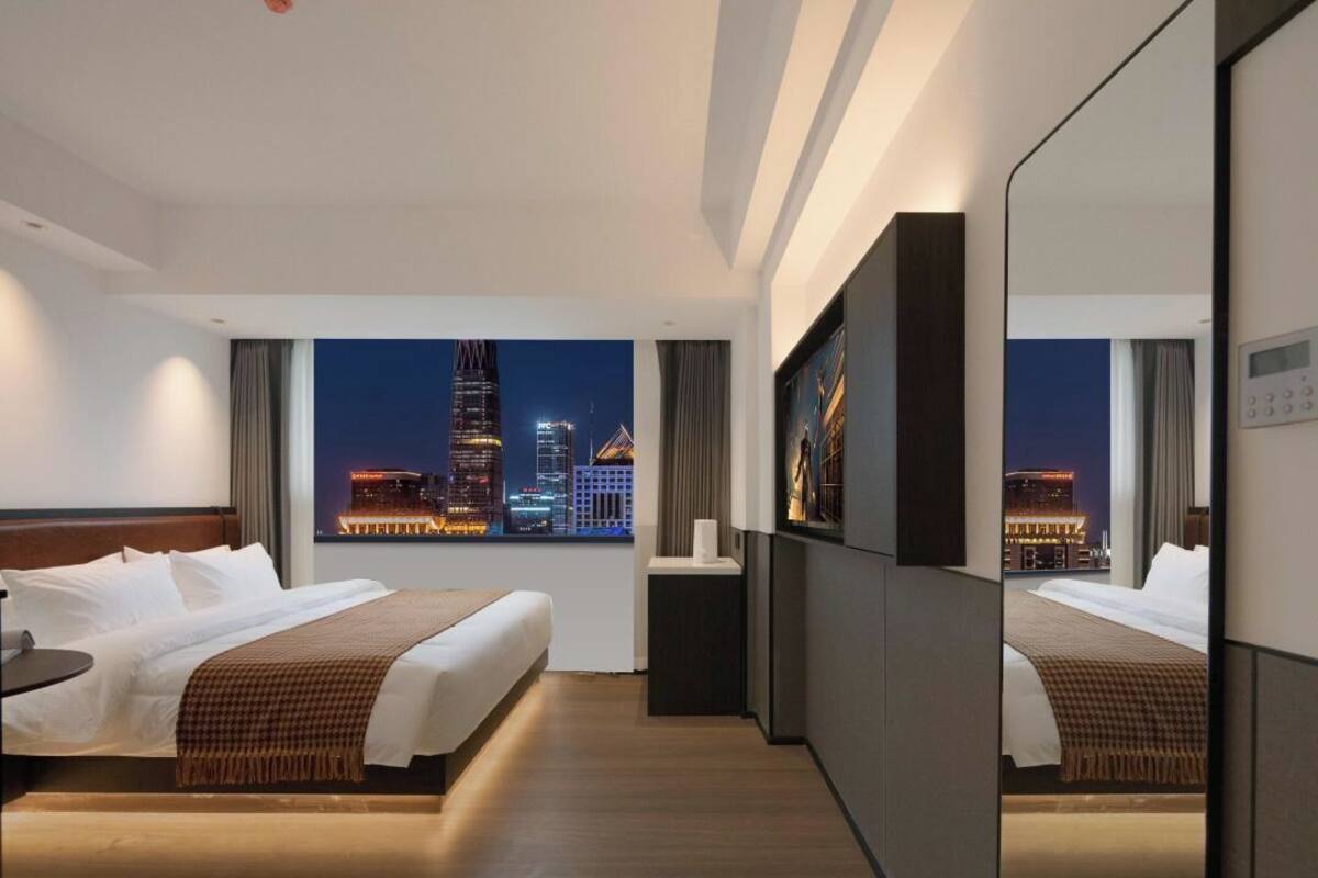 A hotel room with a large bed, a bedside table, a mirror, and a view of a cityscape at night.