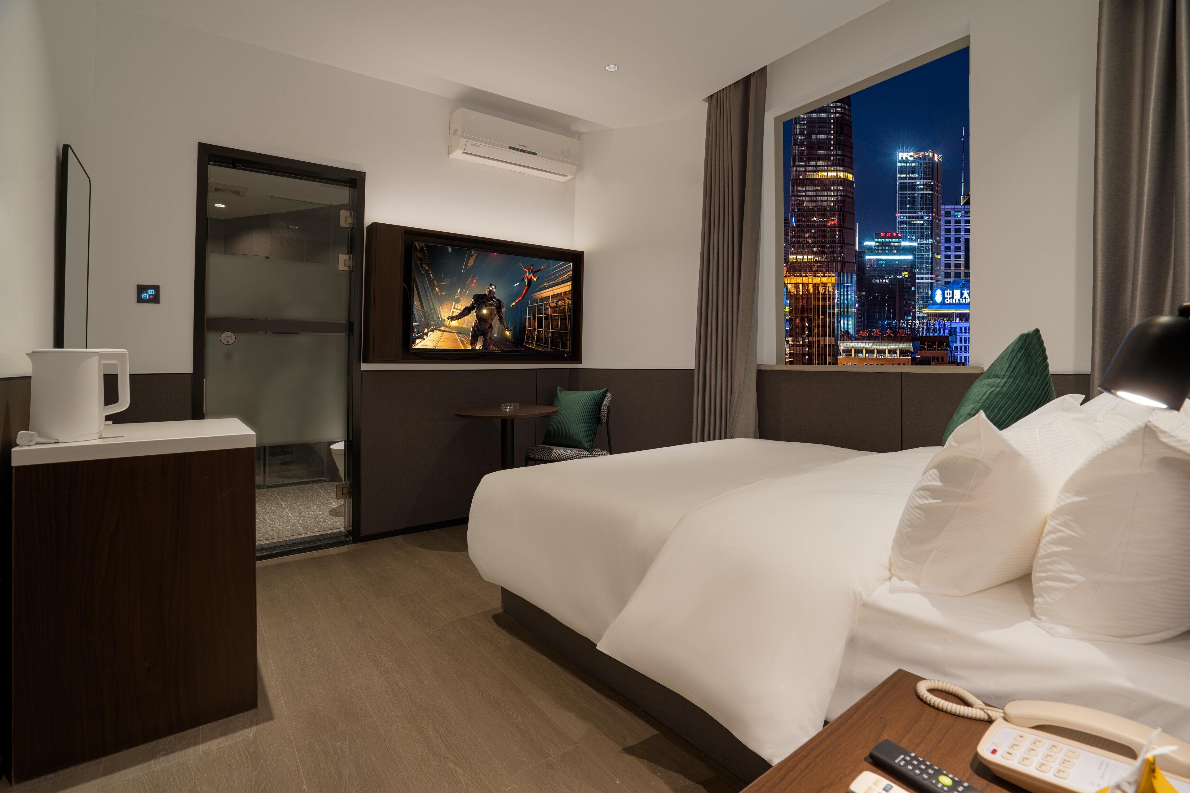 A hotel room with a bed, a desk with a phone, a coffee cup, a painting, a window with a city view, and an air conditioning unit.