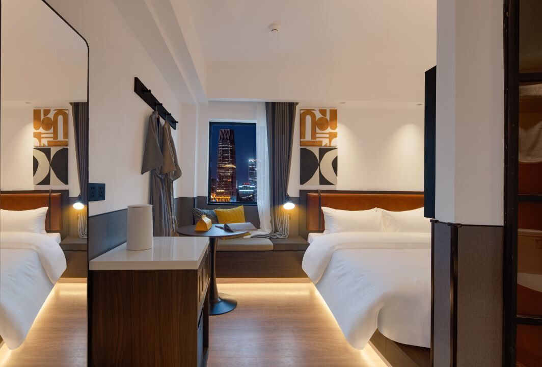 A hotel room with two beds, a desk, and a view of a cityscape at night.