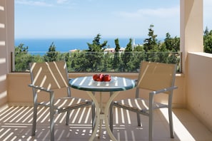 A balcony with two chairs and a table, a bowl of fruit, and a view of the sea and trees. - Blue Hills Villas by Estia Orange 1 (Malevizi)