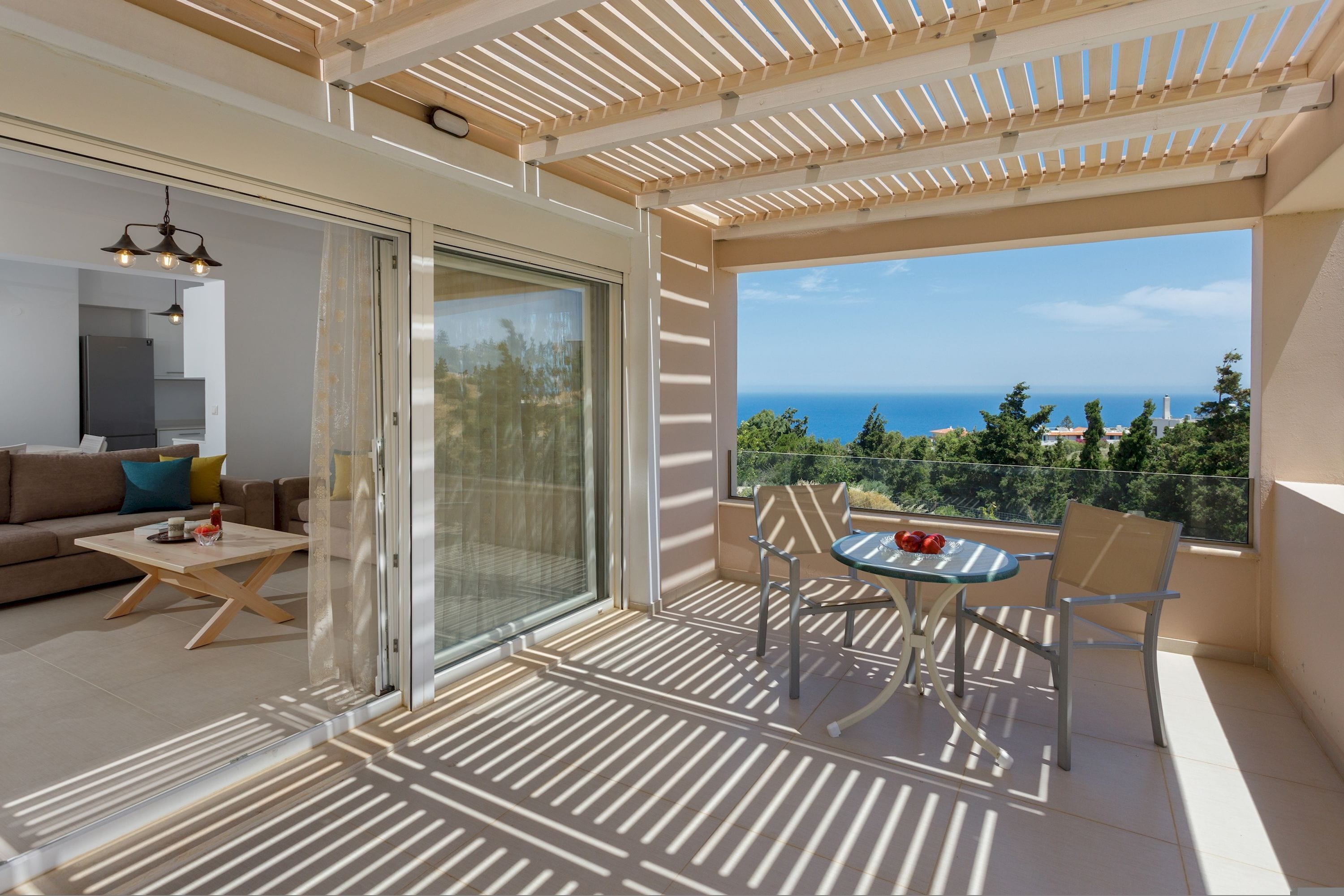 Un balcón con vista al mar, una puerta de vidrio que da a una sala de estar, y una mesa con sillas.