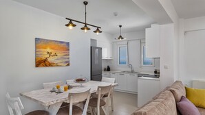 A modern kitchen with white cabinets, a dining table with chairs, and a painting of a sunset. - Blue Hills Villas by Estia Yellow 2 (Malevizi)