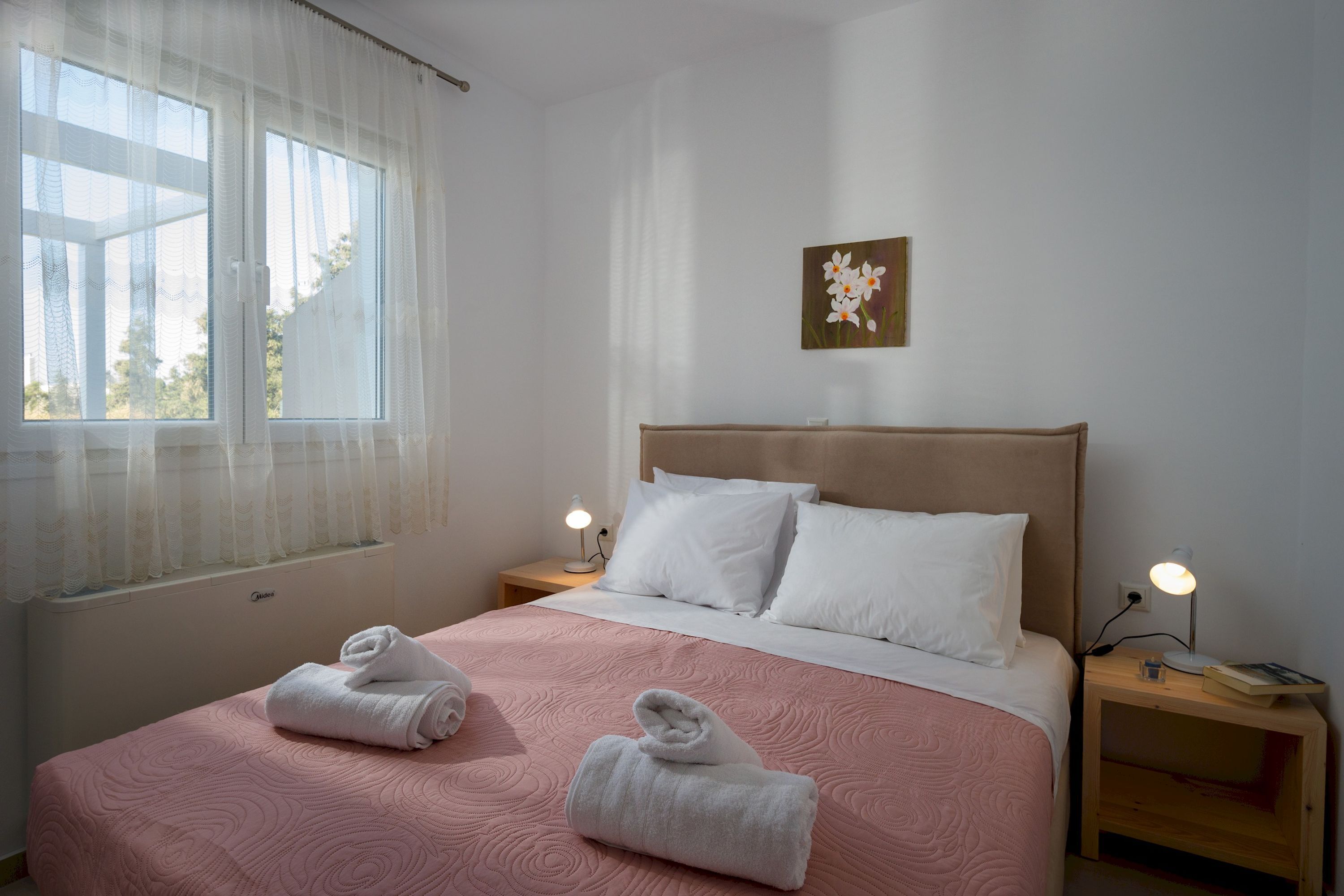 A neatly made bed with pink bedding, two white pillows, and two rolled towels on the bed. There is a nightstand with a lamp on each side of the bed. A window with sheer curtains is visible, and a framed picture hangs on the wall above the bed.