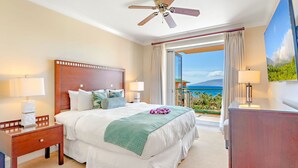 Ein Hotelzimmer mit einem Bett, zwei Nachttischen, einem Deckenventilator, einem großen Fenster mit Vorhängen und Blick aufs Meer.