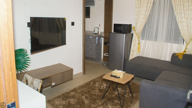 A compact living space with a sofa, a small table, a TV mounted on the wall, a refrigerator, and a kitchen area visible through an open doorway. - 1BD Serenity Dubai Suites (Abuja)