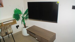 A room with a TV mounted on the wall, a wooden desk, a chair, and a potted plant. - 1BD Serenity Dubai Suites (Abuja)