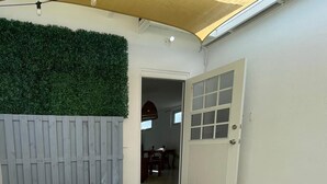 A patio with a white table, a potted plant, a grey fence, and a door leading to an interior space.