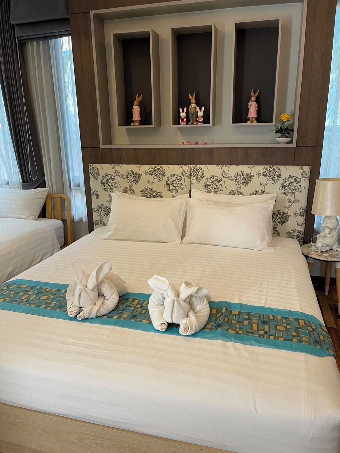 A hotel room with a large bed, two pillows, a headboard with floral pattern, and three small shelves with figurines.