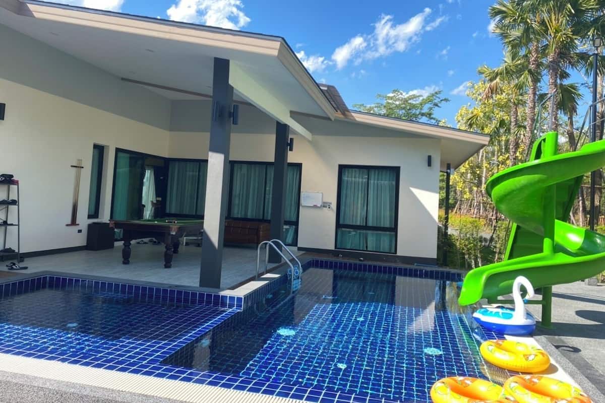 Uma casa moderna com piscina, um escorrega verde e uma área ao redor da piscina com um banco e um piano.