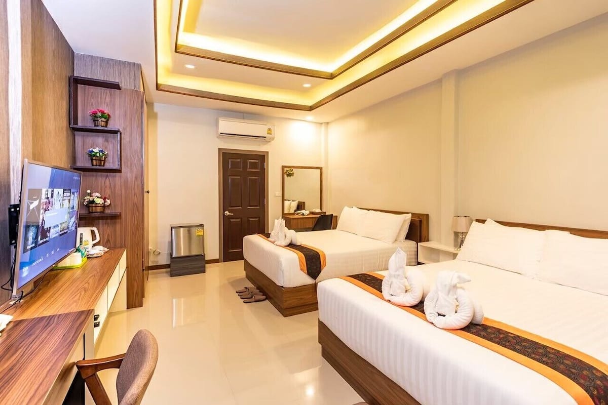 Quarto de hotel com duas camas, uma escrivaninha com computador, televisão e uma prateleira de madeira com objetos decorativos.