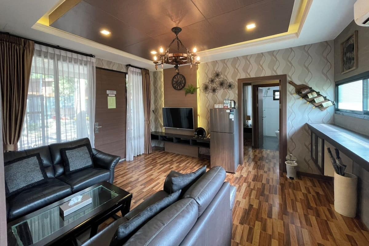Uma sala moderna com sofá, mesa de centro de vidro e televisão. Há uma área de cozinha com frigorífico e uma mesa de jantar. O quarto tem soalho de madeira e uma parede estampada.