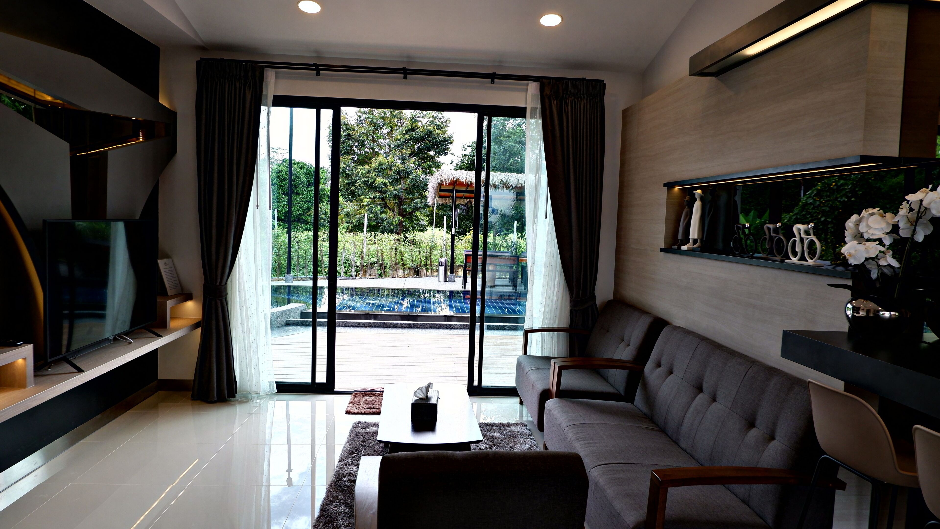 A modern living room with a large sliding glass door leading to a pool area, a flat-screen TV, a grey sofa, and a coffee table.