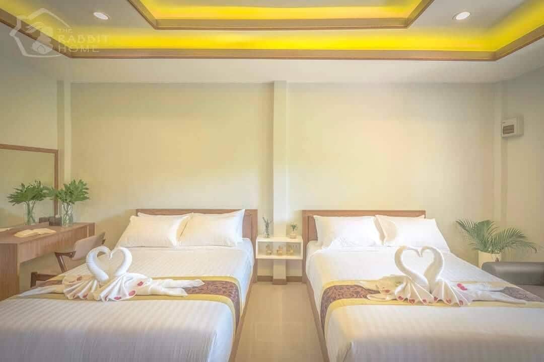 A hotel room with two beds, each with a heart-shaped towel decoration. There is a small table with a lamp and a potted plant.