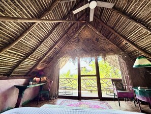 Ein Dachzimmer mit Strohdach, Deckenventilator, großem Fenster und Blick auf Grün.
