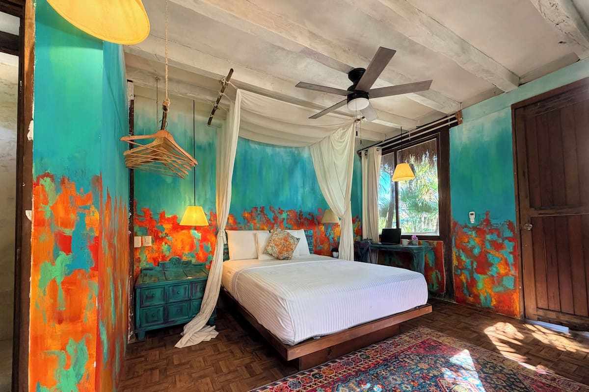 A bedroom with a bed, a ceiling fan, a desk with a lamp, a window, and a door.