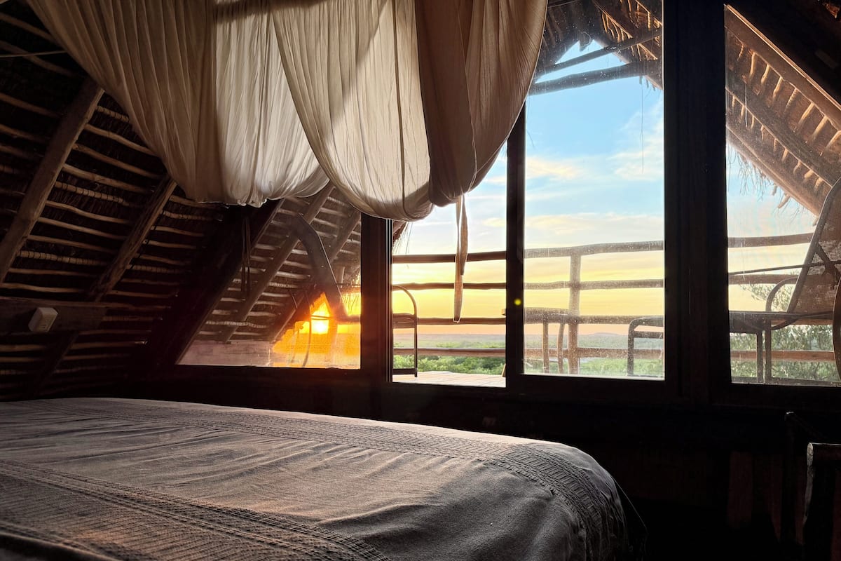 A room with a bed, a window with a view of a sunset, and a thatched roof structure.