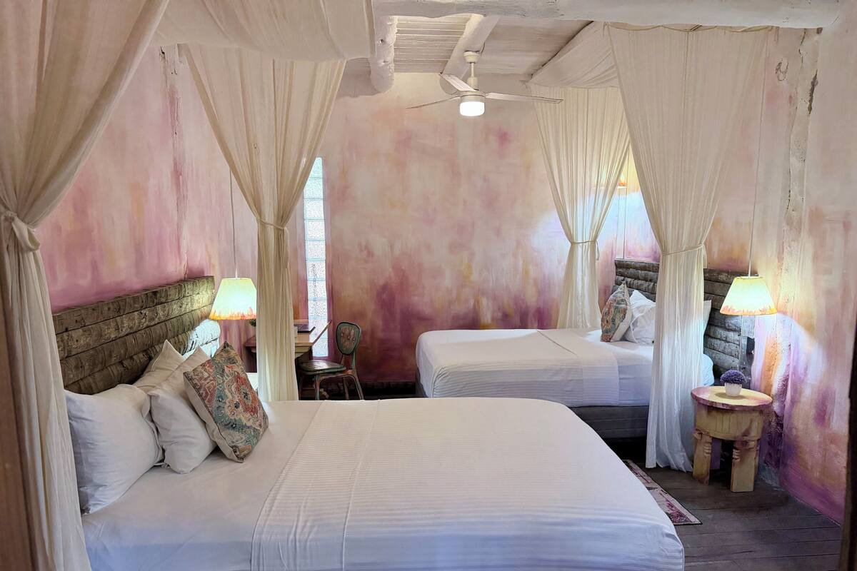 A room with two beds, a wooden headboard, a desk, a chair, and a ceiling fan.