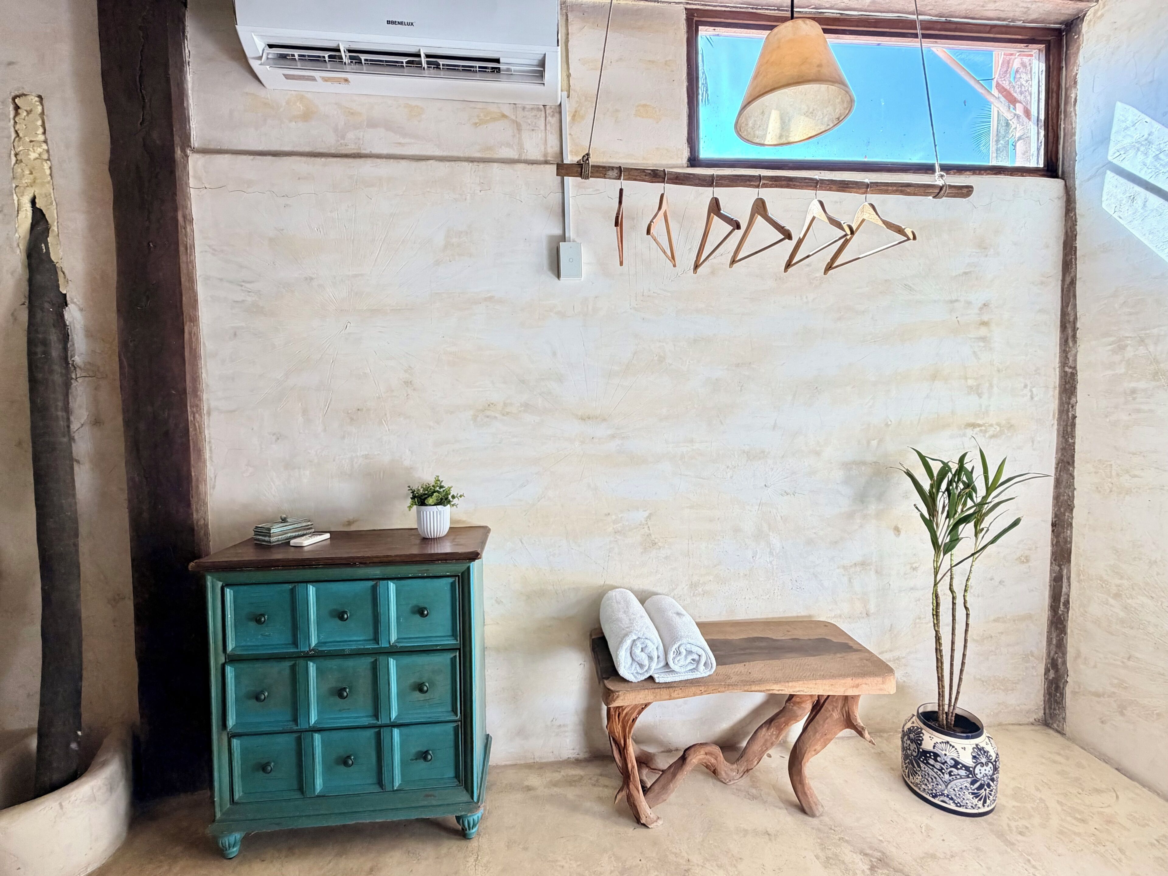 A room with a teal dresser, a wooden bench with towels, a hanging light fixture, and a potted plant.