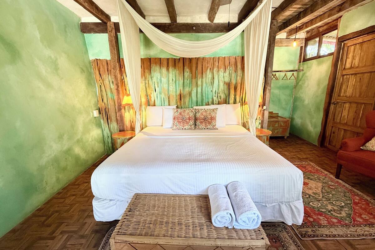 A room with a large bed, a small table with towels, a nightstand, and a wooden door.