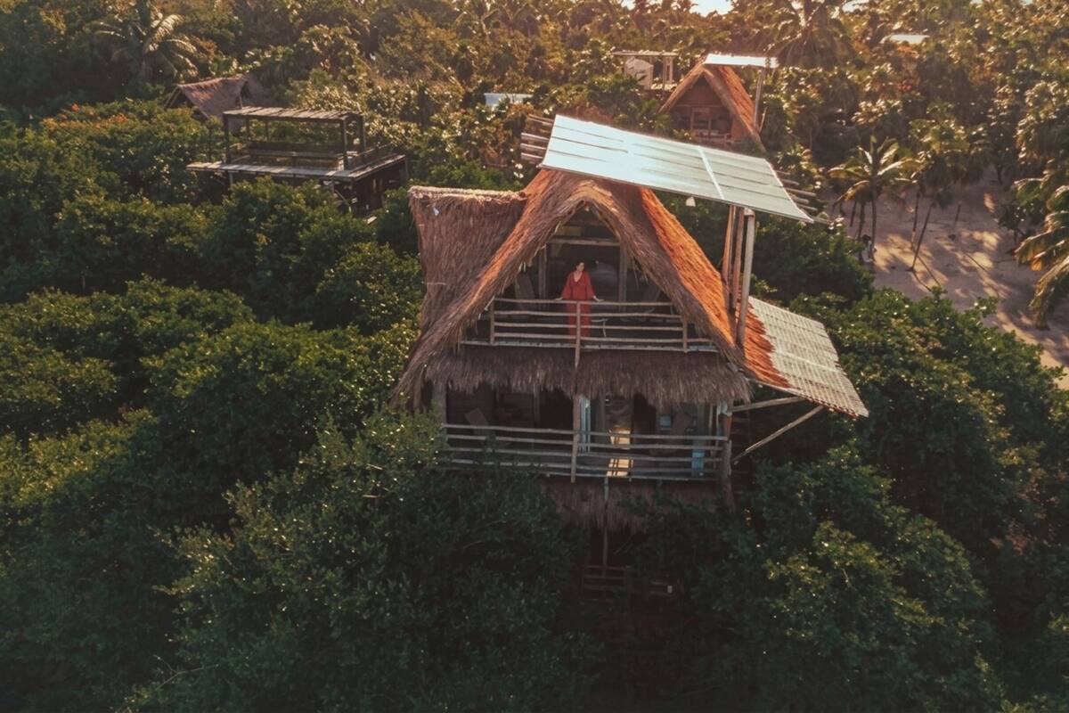 A thatched-roof house with a balcony, surrounded by dense tropical vegetation and a beach in the background.