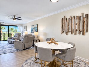 Un salon moderne avec une table à manger ronde, des chaises recouvertes d’un tissu gris, un ventilateur de plafond et une vue sur des palmiers à travers de grandes fenêtres.