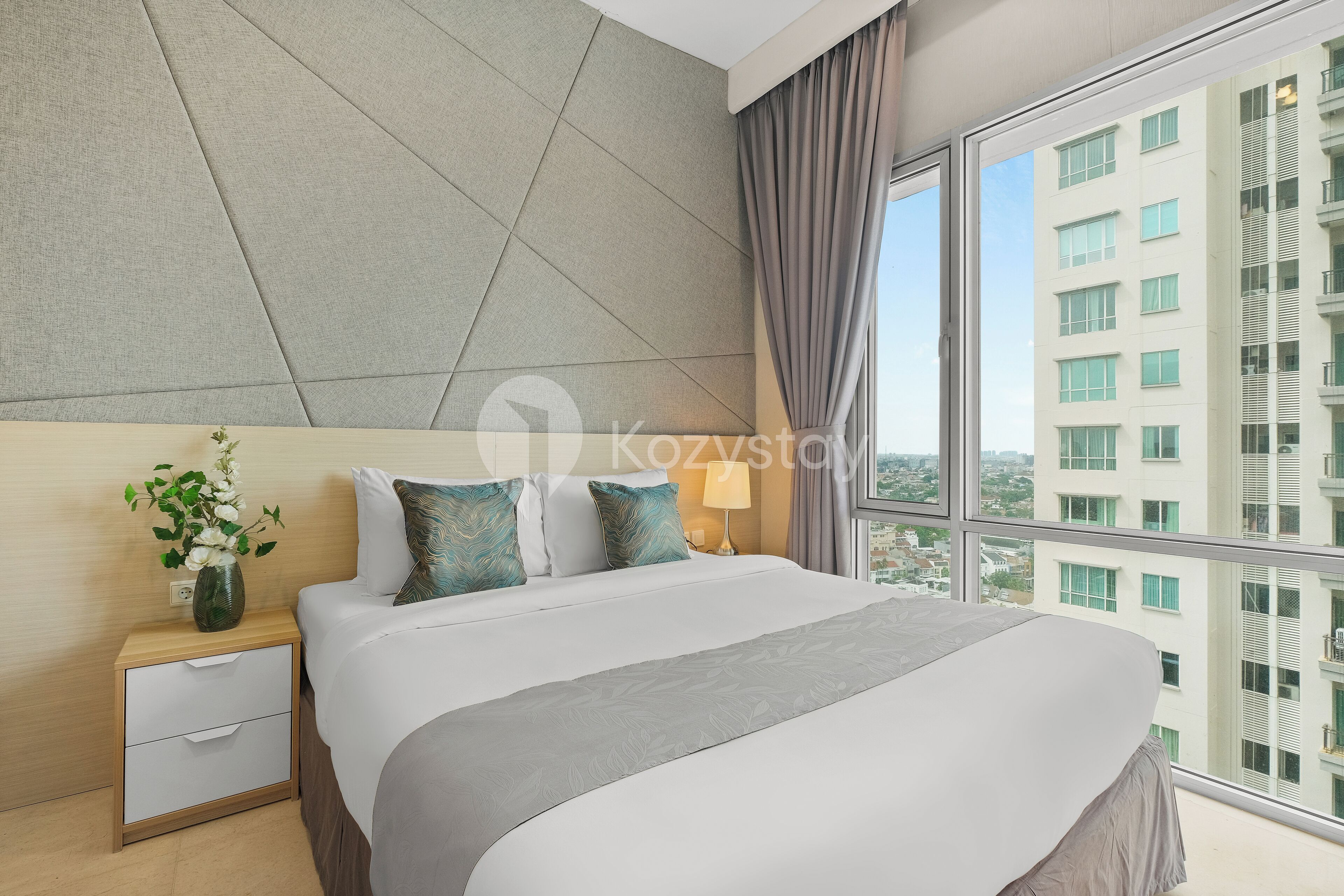 A modern hotel room with a large bed, a nightstand with a vase of flowers, and a view of a cityscape through large windows.