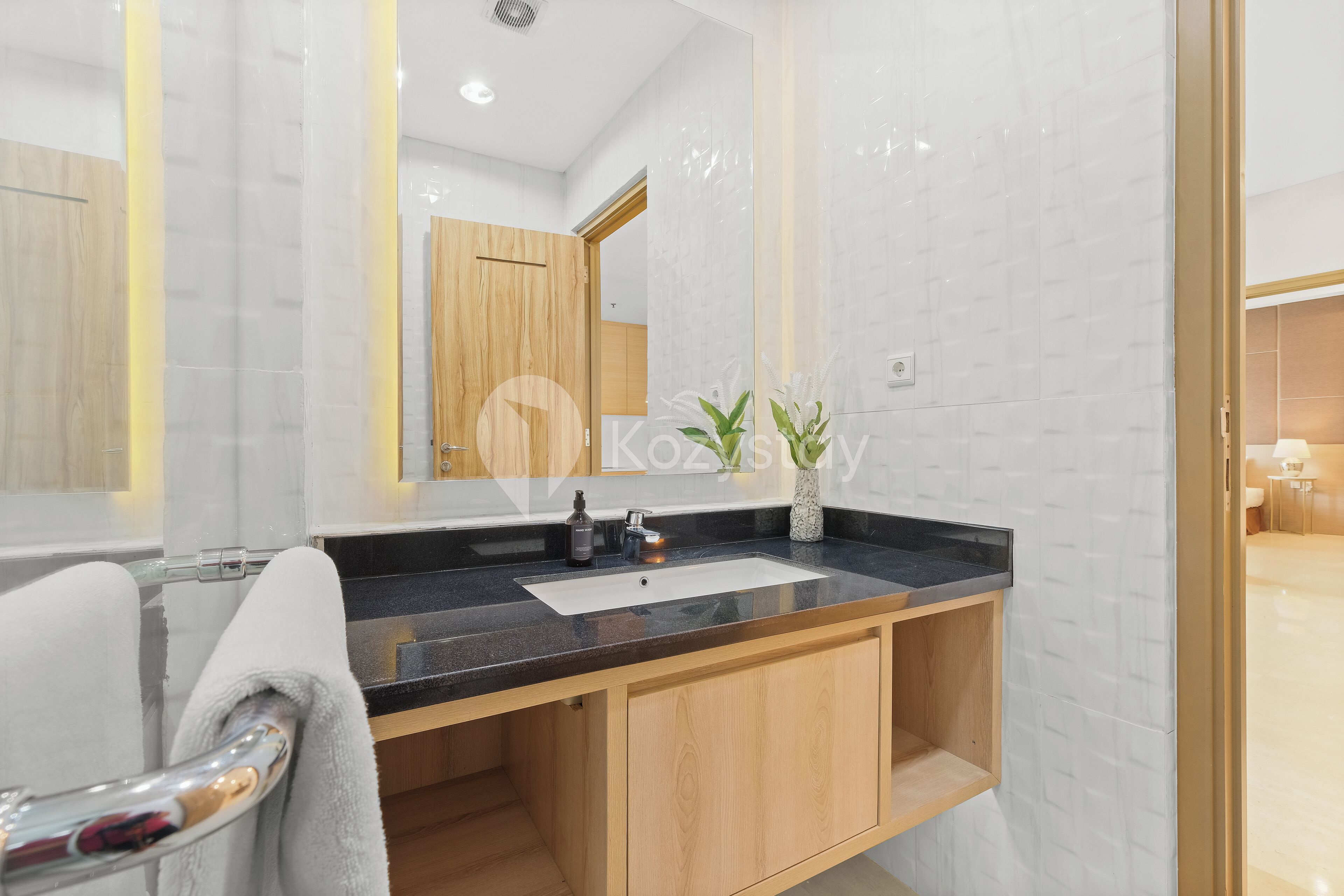 A modern bathroom with a black countertop sink, wooden cabinets, a white towel, and a mirror reflecting a wooden door.